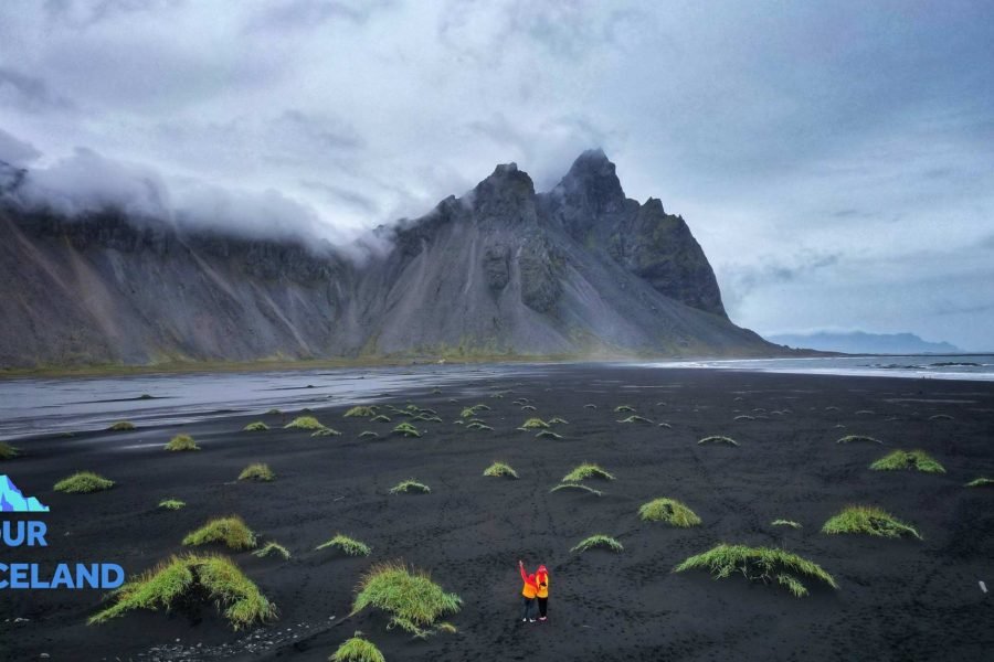 Guests from ouriceland.is posing at the dramatic Black Sand Beach on Iceland’s South Coast during a 4 nights 5 Days Iceland tour package, photographed from a distance with waves and basalt cliffs in the backdrop.